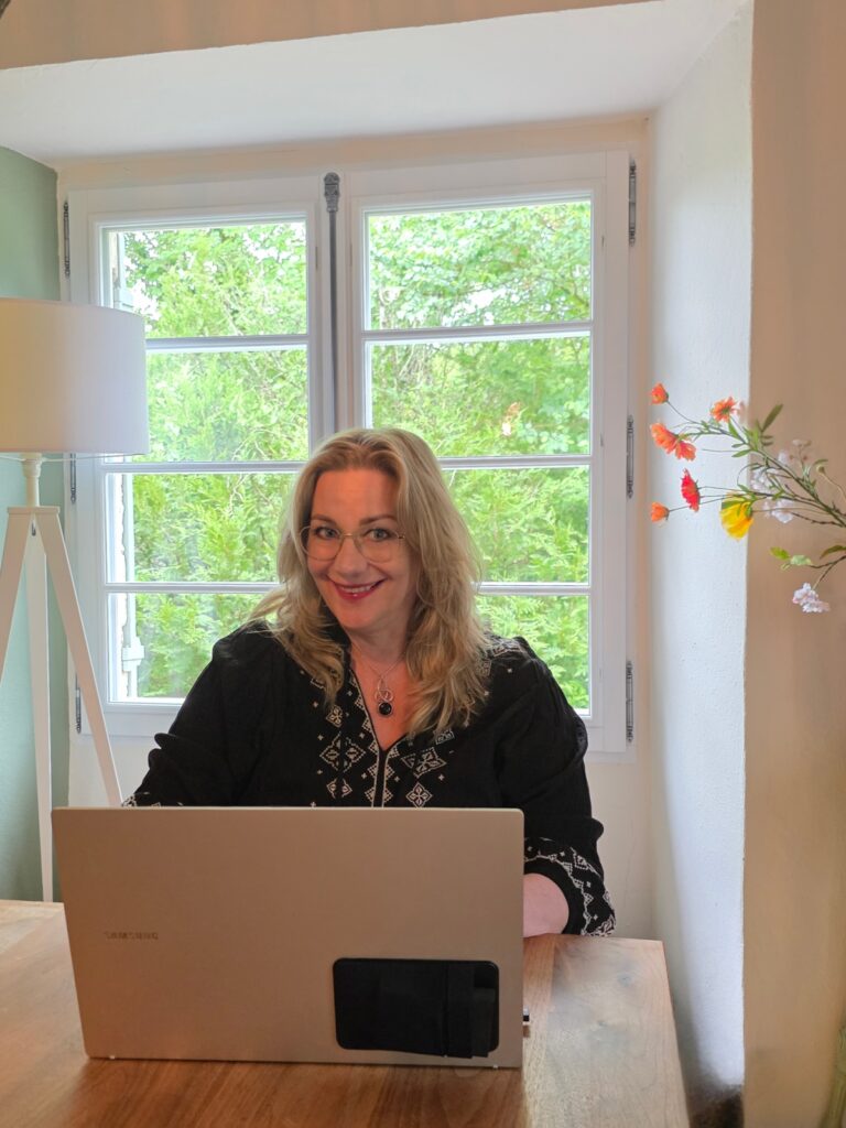 Vrouw met halflang blond haar werkt achter laptop aan houten tafel bij raam met uitzicht op groene bomen