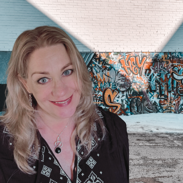 Vrouw met halflang blond haar glimlacht naar de camera, staat buiten voor een muur met kleurrijke graffiti en draagt een zwarte blouse met borduursel